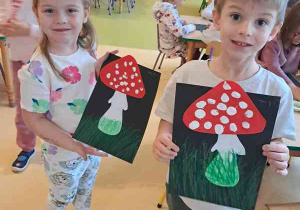 Dziewczynka i chłopiec trzymają w dłoniach wykonane przez siebie prace plastyczne przedstawiające muchomora