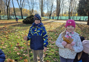 Dziewczynka stoi z bukietem liści a chłopiec z grabiami