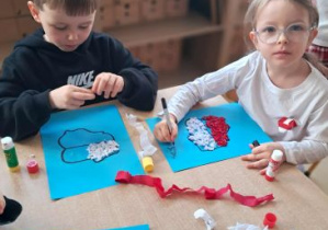 Dzieci siedzą przy stoliku i wyklejają kontury Polski białą i czerwoną bibułą