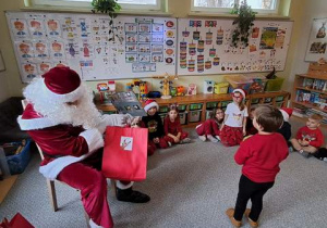 Mikołaj podczas wręczania prezentów, w tle widać dzieci siedzące w kręgu na dywanie
