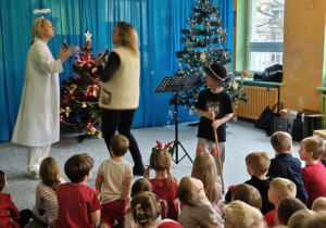 Dzieci siedzą na widowni i słuchają koncertu, na scenie dwie wokalistki oraz chłopiec w czapce góralskiej z ciupagą