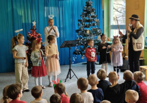 Na scenie stoi wokalistka przebrana za anioła, przed nią stoją dzieci i grają na instrumentach