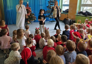 Dzieci siedzą na widowni i słuchają koncertu, na scenie dwie tańczące wokalistki oraz chłopiec w czapce góralskiej z ciupagą