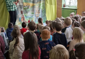 Dzieci siedzą na widowni, na scenie stoi aktor w kraciastej zielonej koszuli i słomianej czapce