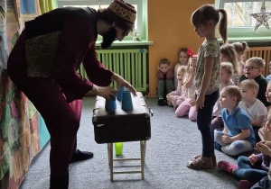 Aktor prezentuje magiczną sztuczkę z kubeczkami ustawionymi na stoliku, dziewczynka stoi przed nim i obserwuje