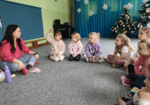 Dzieci siedzą w kręgu, jedna dziewczynka podnosi rękę do góry