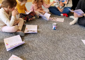 Dzieci siedzą na dywanie, przed nimi leżą książeczki, oglądają zdjęcia pokazywane przez prowadzącą zajęcia