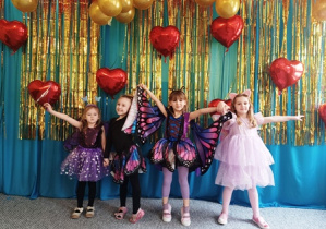 Dzieci ubrane w stroje karnawałowe stoją ustawione na tle niebiesko-złotej kotary przystrojonej balonami