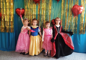 Dzieci ubrane w stroje karnawałowe stoją ustawione na tle niebiesko-złotej kotary przystrojonej balonami