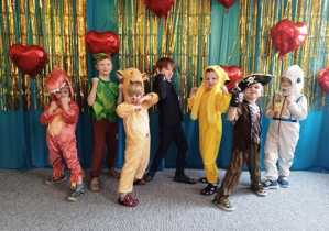 Dzieci ubrane w stroje karnawałowe stoją ustawione na tle niebiesko-złotej kotary przystrojonej balonami