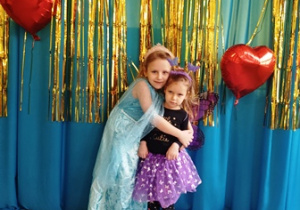 Dzieci ubrane w stroje karnawałowe stoją ustawione na tle niebiesko-złotej kotary przystrojonej balonami