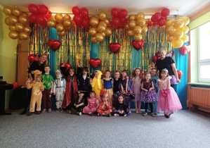 Dzieci ubrane w stroje karnawałowe stoją ustawione na tle niebiesko-złotej kotary przystrojonej balonami