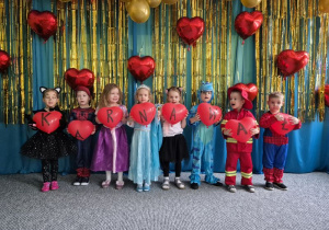 Dzieci ubrane w stroje karnawałowe stoją ustawione na tle niebiesko-złotej kotary przystrojonej balonami, w dłoniach trzymają balony w kształcie serca, które tworzą napis KARNAWAŁ