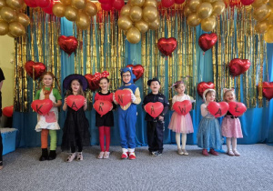 Dzieci ubrane w stroje karnawałowe stoją ustawione na tle niebiesko-złotej kotary przystrojonej balonami, w dłoniach trzymają balony w kształcie serca, które tworzą napis KARNAWAŁ