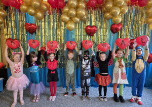Dzieci ubrane w stroje karnawałowe stoją ustawione na tle niebiesko-złotej kotary przystrojonej balonami, w dłoniach trzymają balony w kształcie serca, które tworzą napis KARNAWAŁ