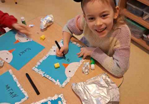 Dziewczynka przy użyciu mazaka podpisuje swoim imieniem pracę plastyczną przedstawiająca bałwanki