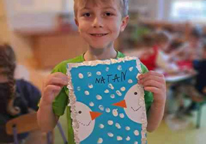 Natan trzyma w dłoniach swoją pracę plastyczną pt. "Bałwanki"