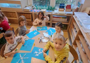 Dzieci w czasie pracy plastycznej wyklejają sylwetę śnieżynki elementami folii aluminiowej