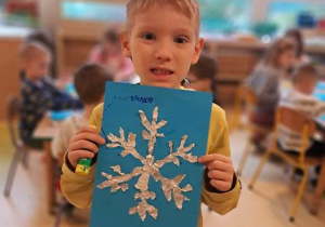 Chłopiec trzyma w dłoniach swoją pracę plastyczną przedstawiającą śnieżynkę wyklejoną z folii aluminiowej