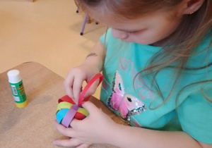 Dziewczynka podczas działań manualnych siedzi przy stole i skleja kolorowe paski papieru tworząc bomkę