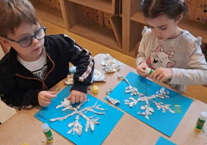 Dziewczynka i chłopiec w czasie wykonywania pracy plastycznej wyklejają sylwetę śnieżynki folią aluminiową