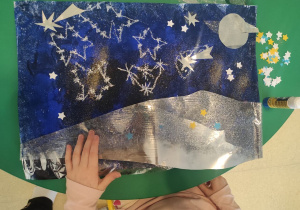 Dziewczynka nakleja folię aluminiową na pracę: ,,Gwiazdozbiory".