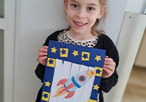 Tosia trzyma w dłoniach swoją kartę pracy przedstawiającą rakietę kosmiczną