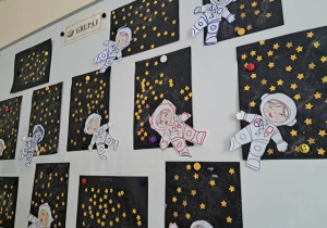 Wystawa prac dzieci z grupy I na tablicy w szatni przedszkolnej prezentująca prace plastyczne pt. "Astronauta w galaktyce"