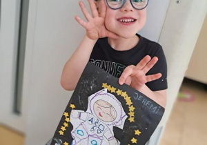 Chłopiec pozuje do zdjęcia i trzyma w dłoniach wykonaną przez siebie pracę plastyczną pt. "Astronauta w galaktyce"