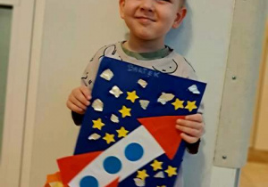 Chłopiec pozuje do zdjęcia i trzyma w dłoniach wykonaną przez siebie pracę plastyczną pt. "Podróż w kosmos"