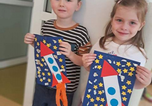 Chłopiec i dziewczynka pozują do zdjęcia i trzymają w dłoniach wykonane przez siebie prace plastyczne pt. "Podróż w kosmos"