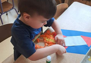 Chłopiec przy stoliku dokleja elementy kolorowego papieru do kartonu tworząc kompozycję rakiety kosmicznej