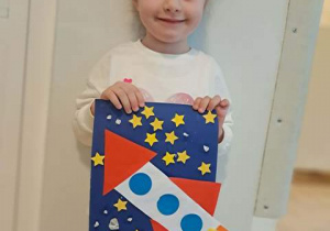 Dziewczynka prezentuje wykonaną przez siebie pracę plastyczną przedstawiającą rakietę kosmiczną