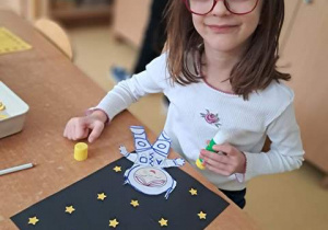 Dziewczynka dokleja gwiazdki z papieru do ciemnego kartonu podczas wykonywania pracy plastycznej pt. "Astronauta w kosmosie"