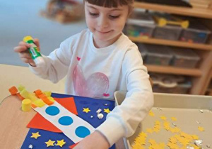 Dziewczynka w czasie wykonywania pracy plastycznej zatytułowanej "Podróż w kosmos" dokleja elementy kolorowego papieru do kartki