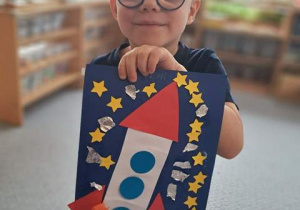 Chłopiec prezentuje wykonaną przez siebie pracę plastyczną przedstawiającą rakietę kosmiczną