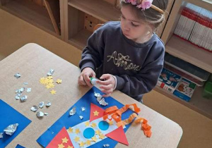 Dziewczyna przy stoliku wykonuje pracę plastyczną przedstawiającą rakietę kosmiczną i dokleja elementy tła wykorzystując folię aluminiową