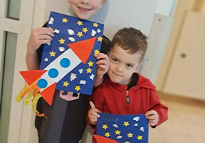 Natan i Ignacy pozują do zdjęcia i trzymają wykonane przez siebie podczas działań plastycznych rakiety kosmiczne