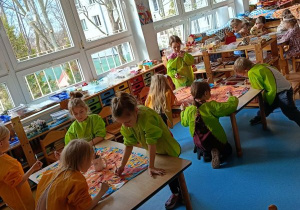 Dzieci stoją dookoła stolików i malują farbami duże kartony w grupach kilkuosobowych