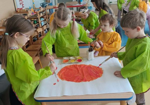 Dzieci stoją dookoła stolika i malują farbami duży karton w grupie kilkuosobowej