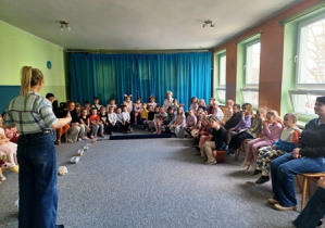 Dzieci siedzą w dużym półkolu słuchając wprowadzenia nauczyciela