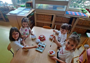 Dziewczynki w czasie działań plastycznych siedzą przy stole i malują brązową farbą kulki uformowane z kartek papieru