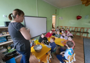 Dzieci siedzą przy stoliczkach i słuchają prowadzącej warsztat