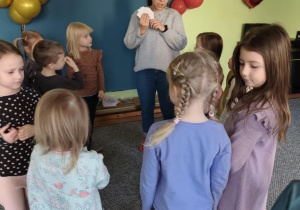 Dzieci stoją w kręgu i słuchają prowadzącej zajęcia, która trzyma w dłoniach białe karteczki