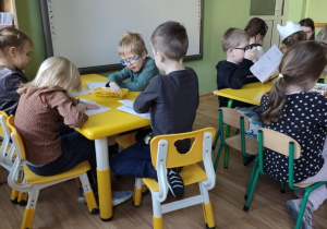 Dzieci siedzą przy stolikach i wykonują rysunki w swoich książeczkach