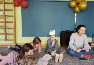 Dzieci leżą na dywanie i oglądają książeczki oraz słuchają opowiadania prowadzącej zajęcia