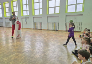 Prowadzący zajęcia trzyma postać człowieka do ćwiczeń, dziewczynka prezentuje prawidłowy sposób wykonania ćwiczenia