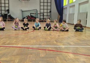 Dzieci siedzą skrzyżnie obok siebie i trzymają piłki w dłoniach