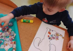 Chłopiec wykleja kolorowymi kawałkami gazety sylwetę psa