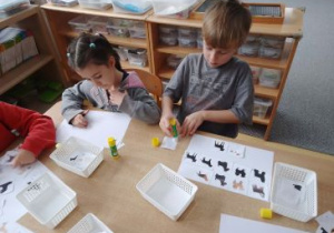 Dzieci siedzą przy stoliku i przyklejają zdjęcia psów oraz dopasowują ich cienie